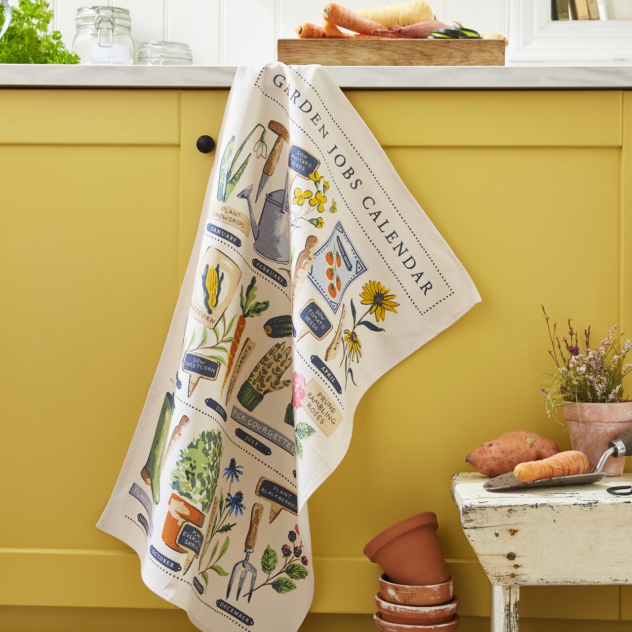 Ulster Weavers Küchentuch im Vintage-Design mit handgemalten Gartenmotiven, wie Gummistiefeln und Pflanzensteckern auf cremefarbenem Hintergrund.